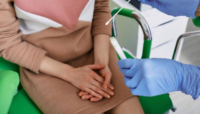 The photograph captures a healthcare provider performing a vaginal exam on a woman sitting on a medical examination table, highlighting the importance of understanding the factors that can affect the balance of vaginal bacteria.


